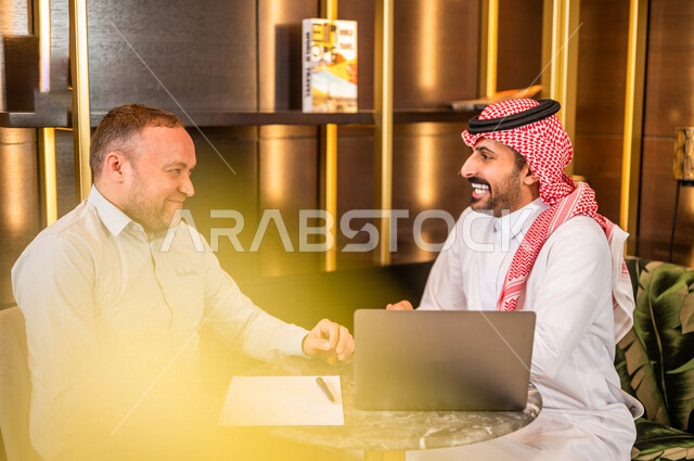 رجلان أعمال عربيان خليجيان سعوديان يستخدمان الحاسوب المحمول و يتناقشان بالعمل ، شركاء عمل يجتمعان في أحد الفنادق الخليجية ، إتفاق عمل ، الشراكة على الأعمال و المشاريع التجارية ، صفقات عمل و تعاون بين الشركات  
