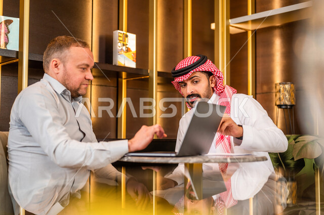 رجلان أعمال عربيان خليجيان سعوديان يستخدمان الحاسوب المحمول و يتناقشان بالعمل ، شركاء عمل يجتمعان في أحد الفنادق الخليجية ، إتفاق عمل ، الشراكة على الأعمال و المشاريع التجارية ، صفقات عمل و تعاون بين الشركات  