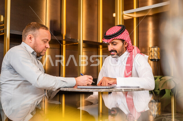 رجل أعمال عربي خليجي سعودي يقوم بتوقيع عقد عمل ، شركاء عمل يجتمعان في أحد الفنادق الخليجية ، إتفاق عمل جديد ، الشراكة على الأعمال و المشاريع التجارية ، صفقات عمل و تعاون بين الشركات