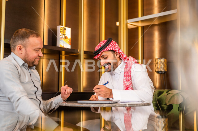 رجل أعمال عربي خليجي سعودي يقوم بتوقيع عقد عمل بسعادة ، شركاء عمل يجتمعان في أحد الفنادق الخليجية ، إتفاق عمل جديد ، الشراكة على الأعمال و المشاريع التجارية ، صفقات عمل و تعاون بين الشركات