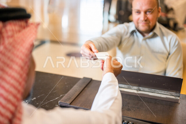 صورة مقربة من الخلف لموظف استقبال عربي خليجي سعودي يقوم بتسليم بطاقة الغرفة الفندقية لسائح عربي ، إجراءات الحجز و الدخول لدى مكتب الاستقبال في الفندق ، خدمة العملاء في الفندق السياحي ، إستلام بطاقة الغرفة ، سياحة و فندقة