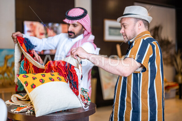 رجل عربي خليجي سعودي يعمل كدليل سياحة برفقة سائح عربي يرتدي لباس الكاجوال في احدي الأماكن التراثية السياحية السعودية ، مشاهدة وسائد المجلس العربي و مطرزات السدو ، مشغولات يدوية ، تراثيات سعودية ، معارض تراثية فنية و شعبية