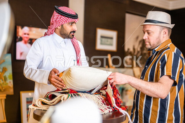 رجل عربي خليجي سعودي يعمل كدليل سياحة برفقة سائح عربي يرتدي لباس الكاجوال في احدي الأماكن التراثية السياحية السعودية ، مشاهدة وسائد المجلس العربي و مطرزات السدو ، مشغولات يدوية ، تراثيات سعودية ، معارض تراثية فنية و شعبية