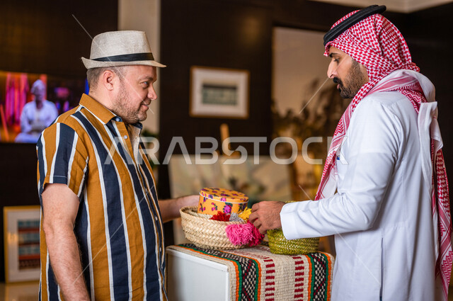 رجل عربي خليجي سعودي يعمل كدليل سياحة برفقة سائح عربي يرتدي لباس الكاجوال في احدي الأماكن التراثية السياحية السعودية ، مشاهدة المطرزات و المشغولات اليدوية ، تراثيات سعودية ، معارض تراثية فنية و شعبية