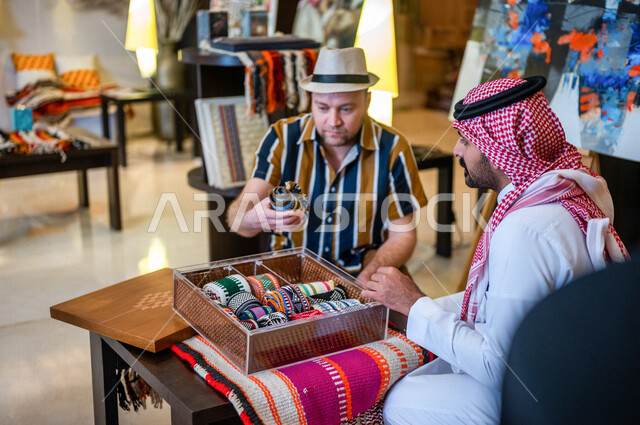 رجل عربي خليجي سعودي يعمل كدليل سياحة برفقة سائح عربي يرتدي لباس الكاجوال في احدي الأماكن التراثية السياحية السعودية ، مشاهدة المطرزات و المشغولات اليدوية ، تراثيات سعودية ، معارض تراثية فنية و شعبية