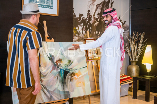 رجل عربي خليجي سعودي يعمل كدليل سياحة برفقة سائح عربي يرتدي لباس الكاجوال في احدي الأماكن التراثية السياحية السعودية ، مشاهدة لوحات فنية تحاكي التراث ، أعمال فنية تشكيلية سعودية ، معارض تراثية فنية و شعبية