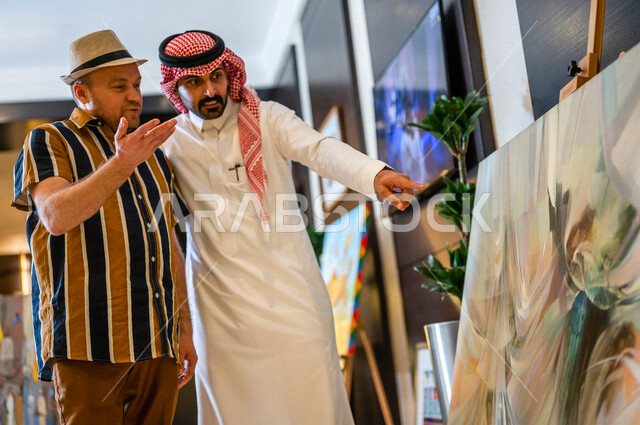 رجل عربي خليجي سعودي يعمل كدليل سياحة برفقة سائح عربي يرتدي لباس الكاجوال في احدي الأماكن التراثية السياحية السعودية ، مشاهدة لوحات فنية تحاكي التراث ، أعمال فنية تشكيلية سعودية ، معارض تراثية فنية و شعبية
