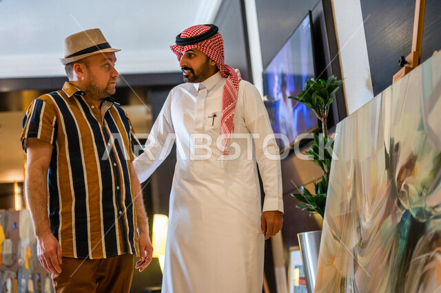 رجل عربي خليجي سعودي يعمل كدليل سياحة برفقة سائح عربي يرتدي لباس الكاجوال في احدي الأماكن التراثية السياحية السعودية ، مشاهدة لوحات فنية تحاكي التراث ، أعمال فنية تشكيلية سعودية ، معارض تراثية فنية و شعبية