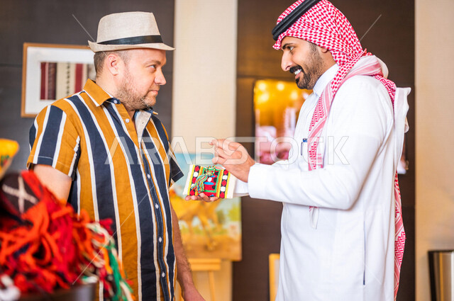 رجل عربي خليجي سعودي يعمل كدليل سياحي برفقة سائح عربي يرتدي لباس الكاجوال مبتسمان في احدي الأماكن التراثية السياحية السعودية ، مشاهدة المطرزات و المشغولات السدو اليدوية ، تراثيات سعودية ، معارض تراثية فنية و شعبية