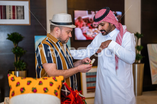 رجل عربي خليجي سعودي يعمل كدليل سياحة برفقة سائح عربي يرتدي لباس الكاجوال في احدي الأماكن التراثية السياحية السعودية ، مشاهدة المطرزات و المشغولات اليدوية ، تراثيات سعودية ، معارض تراثية فنية و شعبية