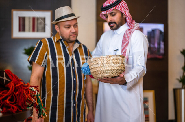 رجل عربي خليجي سعودي يعمل كدليل سياحة برفقة سائح عربي يرتدي لباس الكاجوال في احدي الأماكن التراثية السياحية السعودية ، مشاهدة سلة من سعف النخيل ، صناعات و مشغولات يدوية ، تراثيات سعودية ، معارض تراثية فنية و شعبية