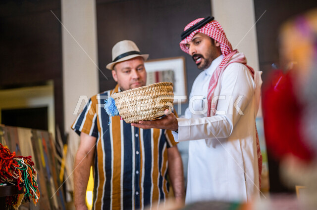 رجل عربي خليجي سعودي يعمل كدليل سياحة برفقة سائح عربي يرتدي لباس الكاجوال في احدي الأماكن التراثية السياحية السعودية ، مشاهدة سلة من سعف النخيل ، صناعات و مشغولات يدوية ، تراثيات سعودية ، معارض تراثية فنية و شعبية