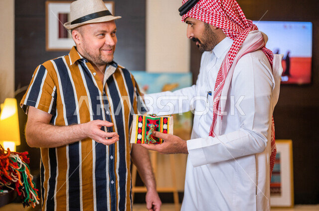 رجل عربي خليجي سعودي يعمل كدليل سياحي برفقة سائح عربي يرتدي لباس الكاجوال مبتسمان في احدي الأماكن التراثية السياحية السعودية ، مشاهدة المطرزات و المشغولات السدو اليدوية ، تراثيات سعودية ، معارض تراثية فنية و شعبية