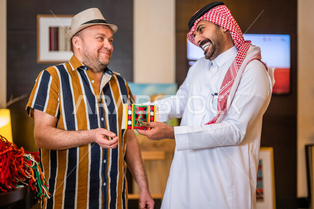 رجل عربي خليجي سعودي يعمل كدليل سياحي برفقة سائح عربي يرتدي لباس الكاجوال مبتسمان في احدي الأماكن التراثية السياحية السعودية ، مشاهدة المطرزات و المشغولات السدو اليدوية ، تراثيات سعودية ، معارض تراثية فنية و شعبية