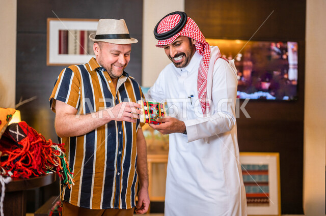 رجل عربي خليجي سعودي يعمل كدليل سياحي برفقة سائح عربي يرتدي لباس الكاجوال مبتسمان في احدي الأماكن التراثية السياحية السعودية ، مشاهدة المطرزات و المشغولات السدو اليدوية ، تراثيات سعودية ، معارض تراثية فنية و شعبية