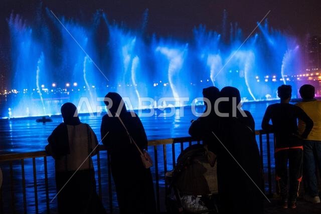 يقف الناس بجانب حاجز لمشاهدة عروض النوافير الملونة وهي تضيء الماء.