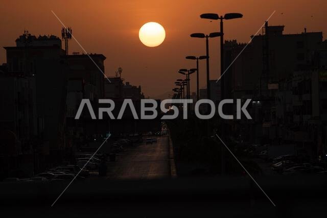 تغرب الشمس الساطعة فوق شارع في المدينة تصطف على جانبيه مبانٍ شاهقة وأعمدة إنارة.