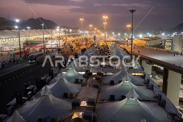 A large number of white tents are set up in a wide outdoor area at dusk, with tall streetlights illuminating the scene and mountains visible in the di