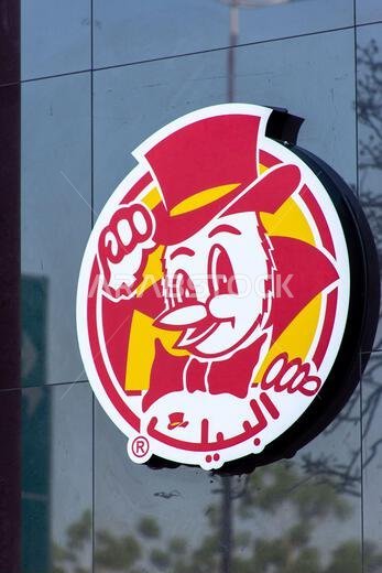 Circular Al Baik fast food restaurant logo sign on a reflective building facade.