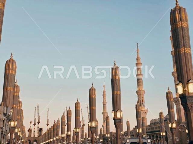 Row of ornate lamp posts leading to tall minarets of Al-Masjid an-Nabawi in Medina, Saudi Arabia, during golden hour with clear sky.