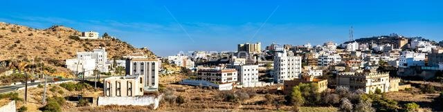 High angle panoramic view of the buildings and mountains in Qarn Dhabi, Al Bahah, Saudi Arabia, showing the urban development and arid hills under a clear blue sky.