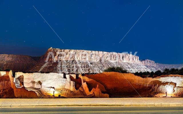 The illuminated ruins of the historic AlUla Old Town at night with a large sandstone mountain and starry sky in the background in Saudi Arabia.