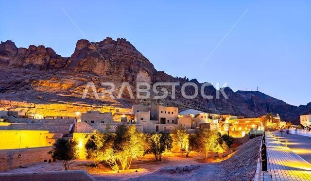 The traditional mud-brick buildings of AlUla Old Town are illuminated at night at the base of a dramatic mountain cliff in Saudi Arabia.