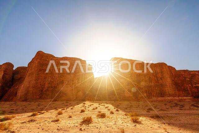 Sun burst creating dramatic light over towering eroded sandstone mountains and arid desert landscape with sparse vegetation in the Sharaan Nature