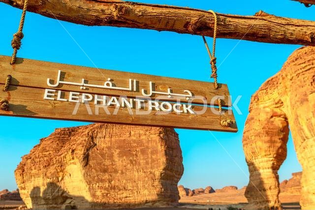 Wooden welcome sign reading Elephant Rock, marks the entrance to the towering sandstone formation overlooking AlUla's desert landscape under blue sky
