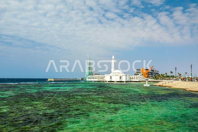 أمواج تتلاطم على شاطئ البحر الأحمر، ومسجد الرحمة الأبيض، ومبانٍ حديثة ترتفع على طول كورنيش جدة في المملكة العربية السعودية
