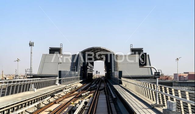 Detailed view of the futuristic Riyadh Metro Al Hamra Station with railway tracks leading into the facility under a clear blue sky in Saudi Arabia.