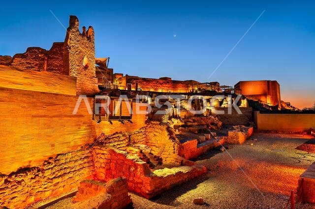 The historic mud brick ruins of At Turaif in Diriyah, Saudi Arabia, illuminated at dusk under a blue twilight sky.