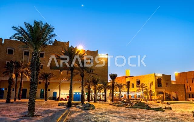 المباني التقليدية ذات الطراز النجدي وأشجار النخيل في ساحة سوق الزال مضاءة ليلاً في الرياض، المملكة العربية السعودية.