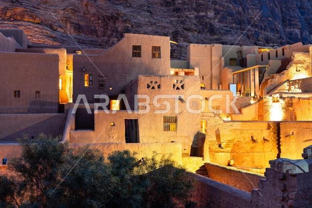 View of the illuminated historic mud brick architecture in the AlUla Old Town heritage site at dusk with rocky mountains in the background, Saudi Arabia.