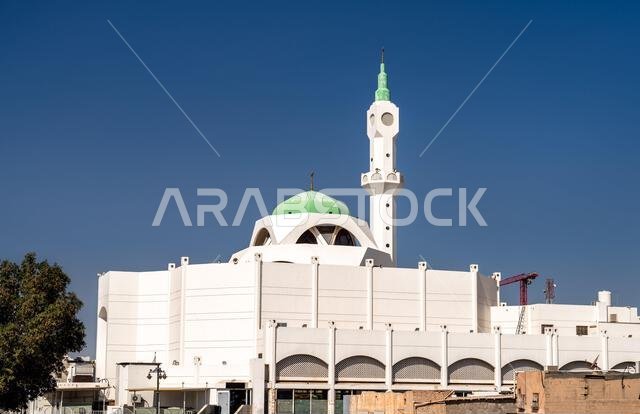Exterior view of the Bilal Ibn Rabah Mosque with its green dome and minaret under a clear blue sky in Madinah, Saudi Arabia