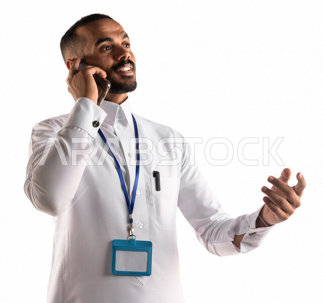 Portrait of a Saudi Arabian Gulf man wearing a white blank identification card, using a mobile phone to make a phone call, a work call, communication and social communication, following up on work goals and tasks, white background