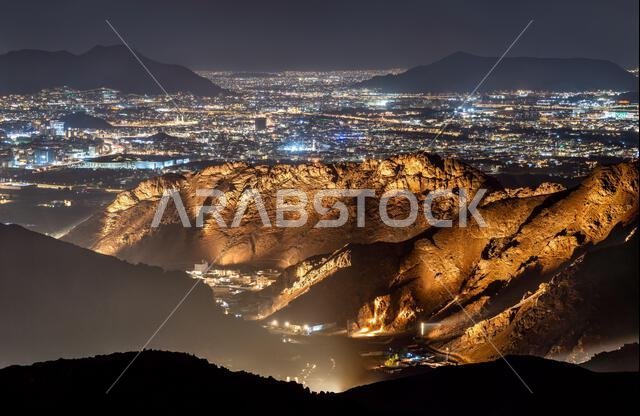 A panoramic night view from Mount Uhud overlooking the illuminated cityscape of Madinah with rugged mountain ridges in the foreground.
