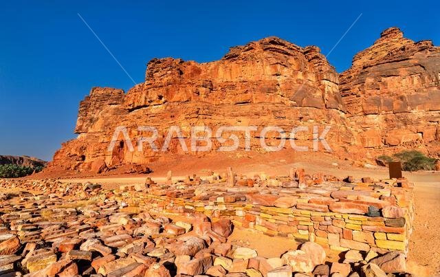 Ancient stone ruins at the Dadan archaeological site are set against massive red sandstone cliffs in AlUla, Saudi Arabia.