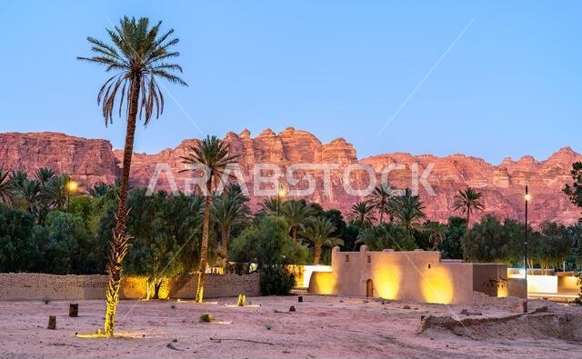 منظر مسائي خلاب للعمارة التقليدية المبنية من الطوب اللبن وبستان النخيل المورق في مدينة العلا القديمة مقابل جبال الحجر الرملي الأحمر في المملكة العربية السعودية.