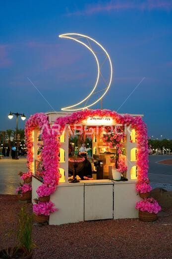 Decorated food kiosk with pink flowers and an illuminated neon crescent moon at blue hour in Madinah, Saudi Arabia