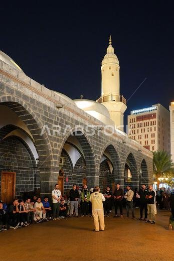 مرشد سياحي يتحدث إلى الحجاج تحت الأقواس الحجرية لمسجد الغمامة ليلاً في المدينة المنورة، المملكة العربية السعودية