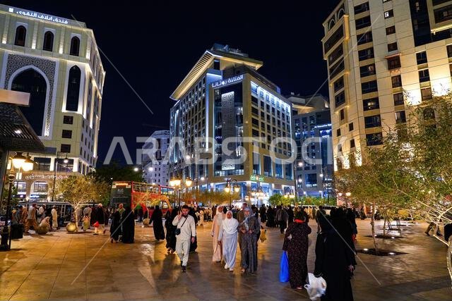 حجاج يسيرون ليلاً في ساحة المناخة المضاءة بالقرب من الفنادق الفاخرة في المدينة المنورة، المملكة العربية السعودية
