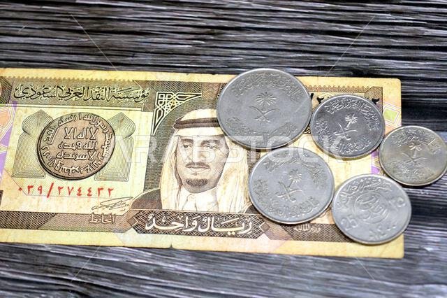 عملات معدنية سعودية قديمة بقيم مختلفة على ورقة نقدية سعودية، منها ريال واحد، وبيانات تاريخية، وسعر صرف الريال السعودي، وإحصاءات الاقتصاد.