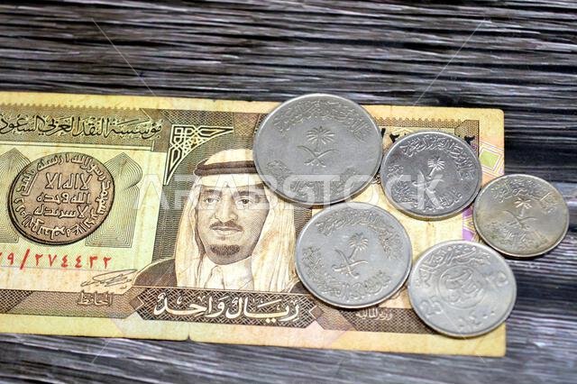 عملات معدنية سعودية قديمة بقيم مختلفة على ورقة نقدية سعودية، منها ريال واحد، وبيانات تاريخية، وسعر صرف الريال السعودي، وإحصاءات الاقتصاد.