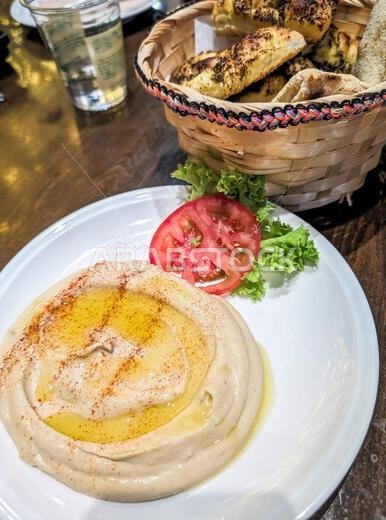 حمص تقليدي مغطى بزيت الزيتون، يقدم مع سلة من الخبز الطازج على طاولة في مطعم محلي في الرياض، المملكة العربية السعودية
