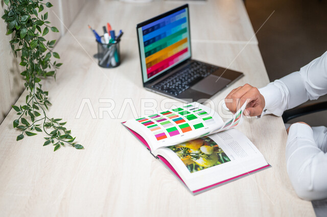 صورة مقربة ليد مصمم عربي خليجي سعودي يجلس على مكتب العمل ، يستخدم الحاسوب المحمول لعرض مخطط الألوان ، كتاب أساسيات الألوان ، تصميم الرسومات و نظرية الألوان ، انتقاء الألوان و اختيارها بالتصميم ، أدوات لوحات ومخططات الألوان