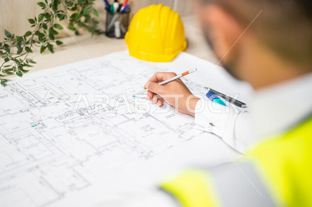 صورة مقربة من الخلف لمهندس عربي خليجي سعودي داخل مكتب هندسي ، يقوم بإستخدام أدوات و معدات هندسية على مخطط تصميم هندسي ، متابعة المشروع و تدقيق العمل ، خوذة وقاية ، مهن و وظائف هندسية ، مهندس معماري محترف ، مفهوم الهندسة المعمارية 