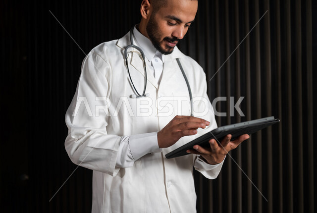 بورتريه لطبيب عربي خليجي سعودي يرتدي المعطف الطبي و السماعة الطبية ، يحمل جهاز التابلت ، كتابة تقرير طبي لحالة مرضية ، تقديم الخدمات الطبية للمرضى ، تقديم الاستشارات الطبية عبر الانترنت ، وظائف طبية