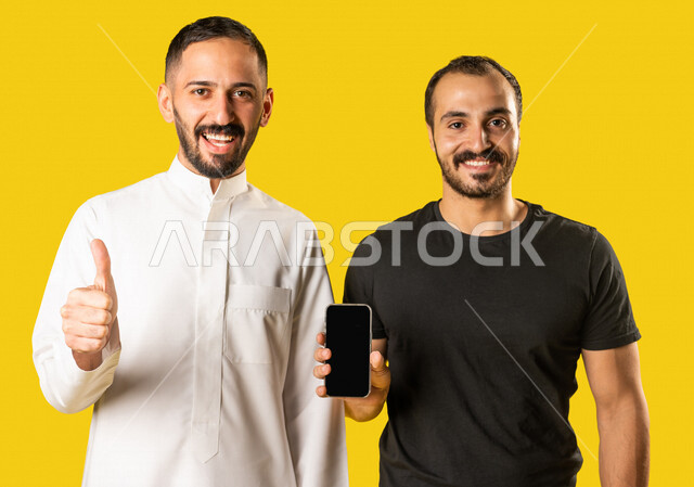 بورتريه لرجلان عربيان خليجيان سعوديان، احدهم يمسك بيده الهاتف المحمول والاخر يرفع اصبع الابهام للاعلى، شاشة سوداء فارغة، عرض خدمات وتطبيقات الهاتف المحمول، التسوق الإلكتروني، ايماءات وجه ودين تدل على الاعجاب والقبول، خلفية صفراء