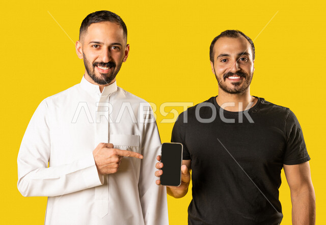 بورتريه لرجلان عربيان خليجيان سعوديان، احدهم يمسك بيده الهاتف المحمول والاخر يشير اليه باصبعه السبابة، عرض شاشة سوداء، أجهزة تقنية حديثة، شاشة سوداء فارغة، عرض خدمات وتطبيقات الهاتف المحمول، التسوق الإلكتروني، مفهوم الإعلانات، خلفية صفراء